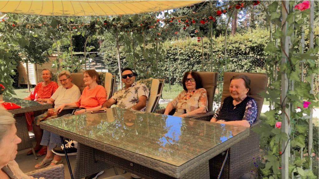 Bewohner der Wohngemeinschaft sitzen mit Gästen aus Tagespflege am Tisch im Garten